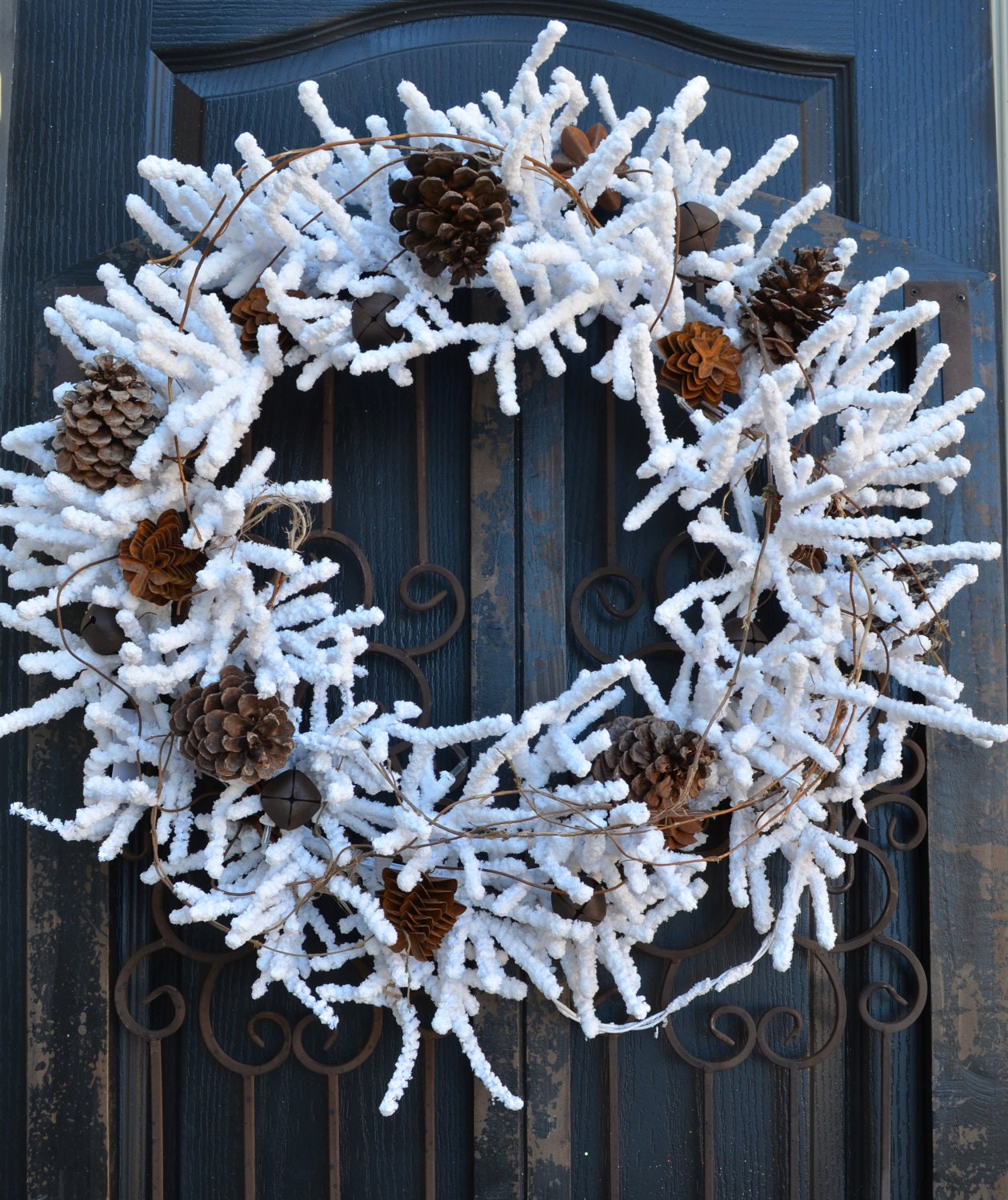 White flocked wreath lighted with rusted pinecones, natural pinecones and rusted jingle bells. - DoorBling