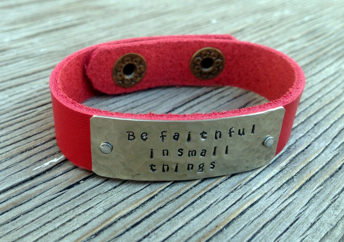 A riveted, 1/2 inch wide red leather cuff bracelet hand stamped with be faithful in small things